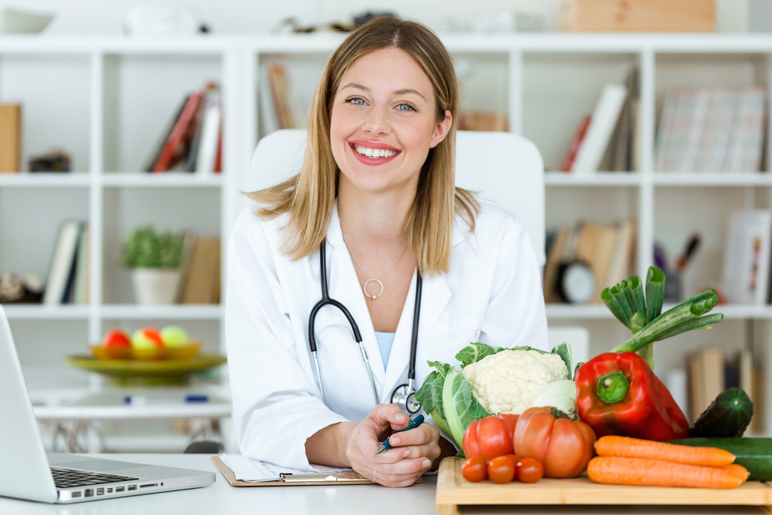 Beautiful smiling nutritionist looking at camera and showing healthy vegetables in the consultation.